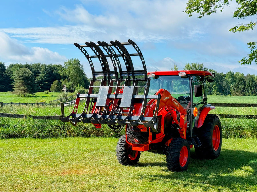A compact tractor with a hydraulic grapple on its front loader, the curved steel jaws held open above a field