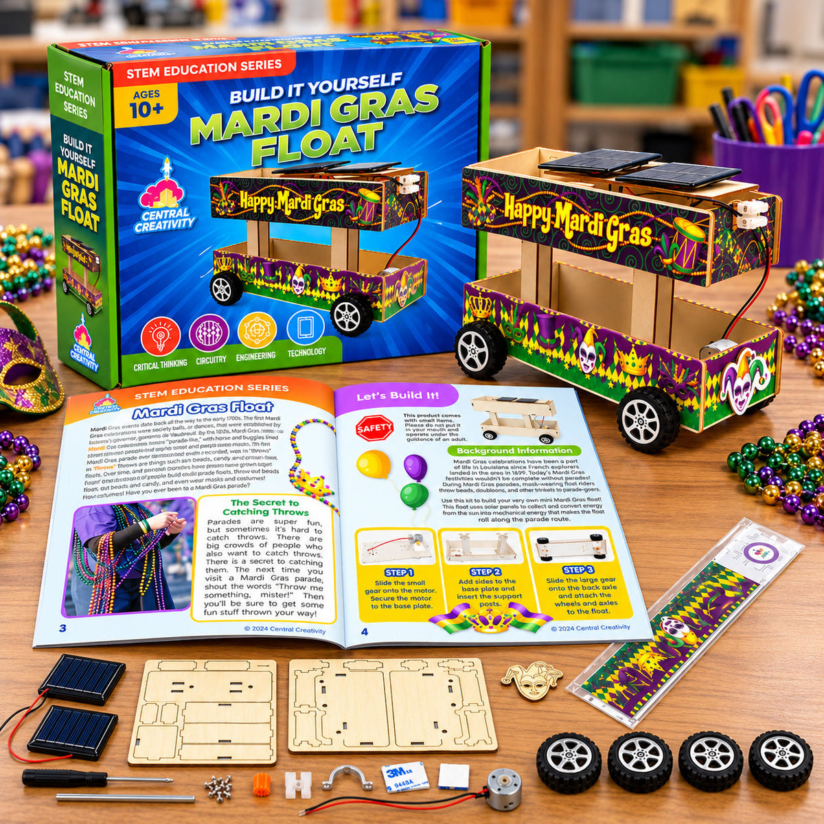 The Mardi Gras Float kit laid out on a classroom table: the printed box, the finished float with its solar panels, the instruction manual open to a build spread, and the parts set out below them, including two solar panels, laser-cut wooden sheets, four wheels, gears, a motor, axles, a screwdriver and a sheet of decorative wraps.