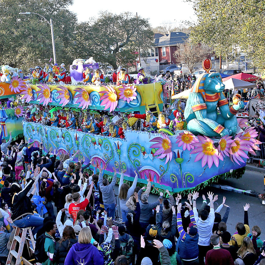 A two-deck Mardi Gras parade float passing a crowd with their hands in the air. Costumed, masked riders line both decks throwing beads; the side panels are airbrushed with pink lotus flowers and green scrollwork over pale blue, a green fringe skirt hides the trailer below, and a turquoise sculpted baboon figure taller than the riders sits at the back end.