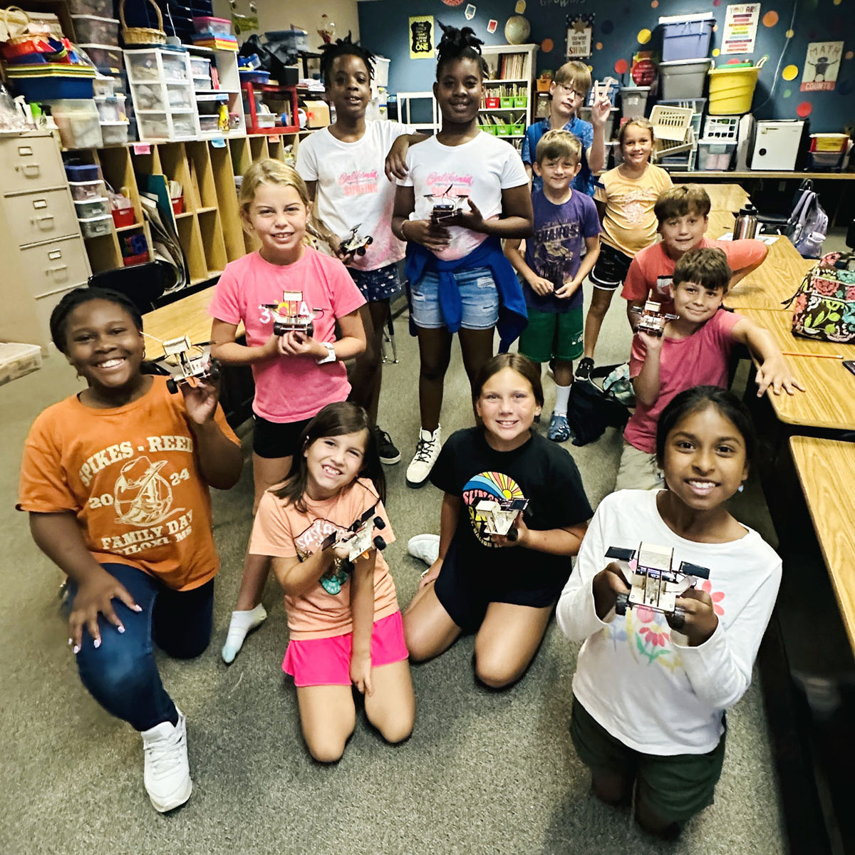About a dozen campers crowded together for a photograph in a busy makerspace, nearly every one of them holding up the small cardboard machine they built, all of them grinning at the camera.