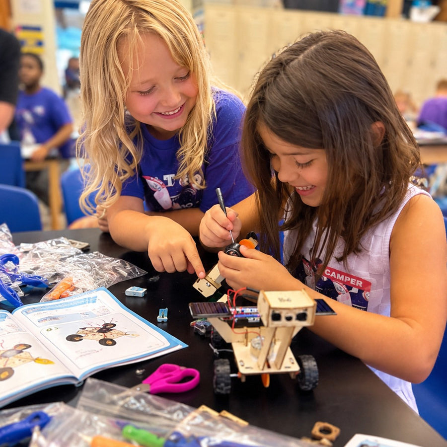 Two girls lean over a camp table building a wooden motorized rover fitted with a small solar panel, one holding it steady while the other turns a screwdriver, an open illustrated guide and bags of parts spread around them and other campers in blue camp shirts at the tables behind.