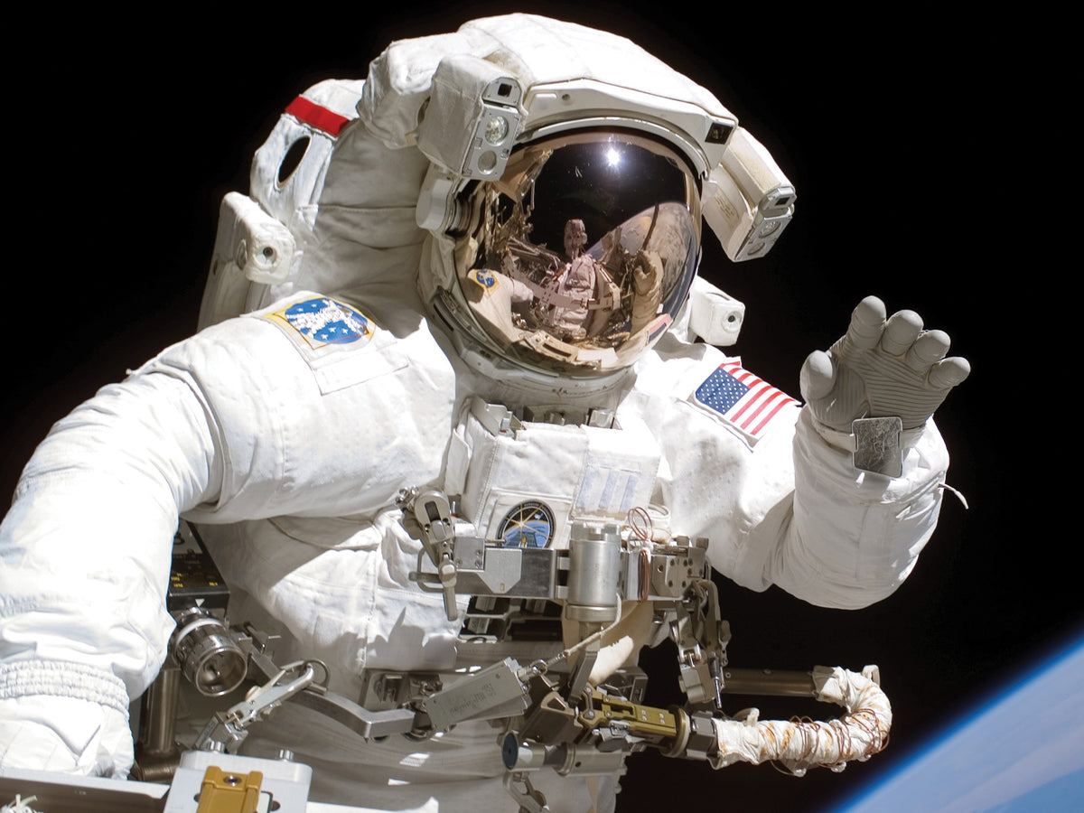 A NASA astronaut on a spacewalk in a white spacesuit, one gloved hand raised, the gold visor reflecting the space station, with the blue curve of Earth below against black space