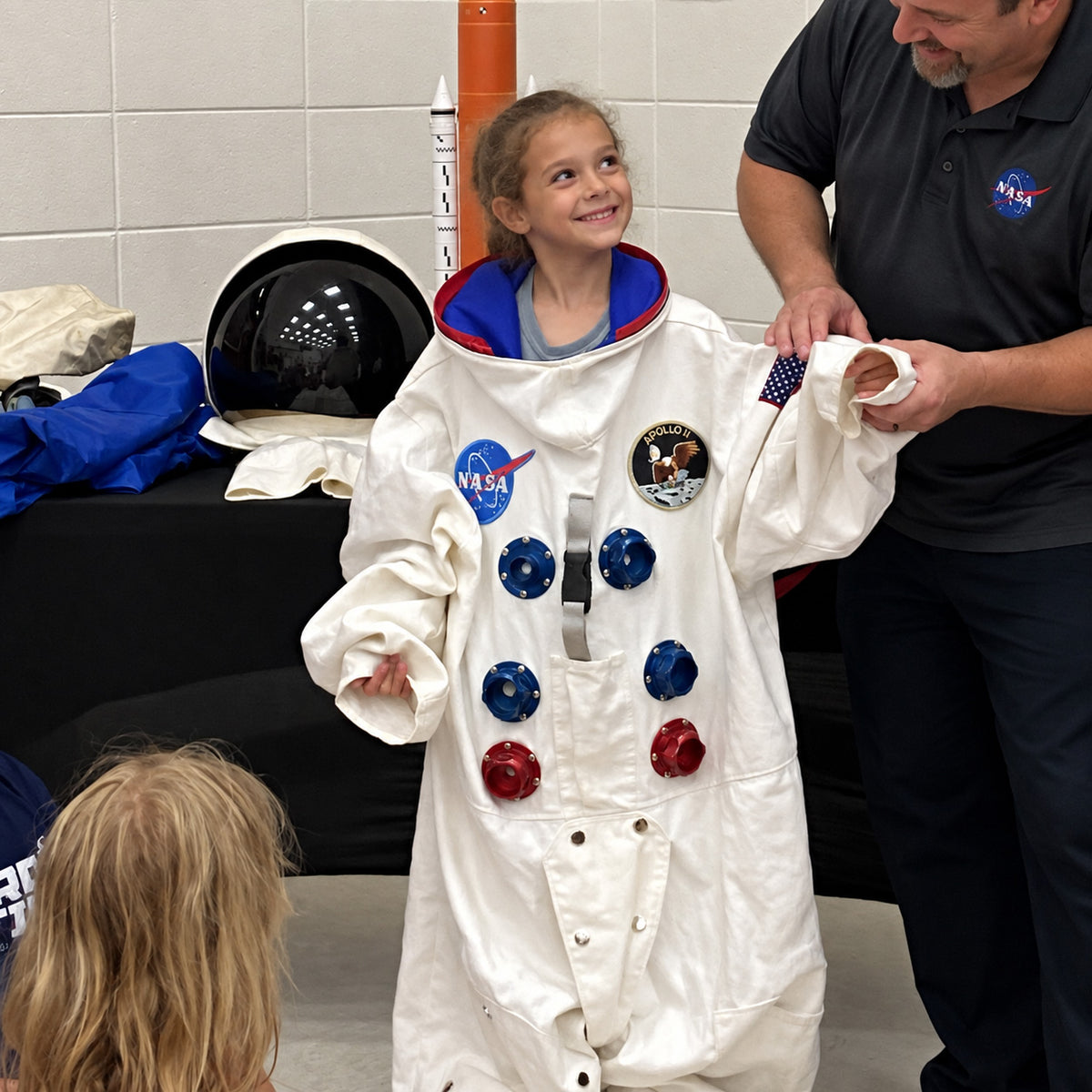 A girl grinning up at a NASA staff member as he helps her into a full white Apollo-era spacesuit jacket, several sizes too big