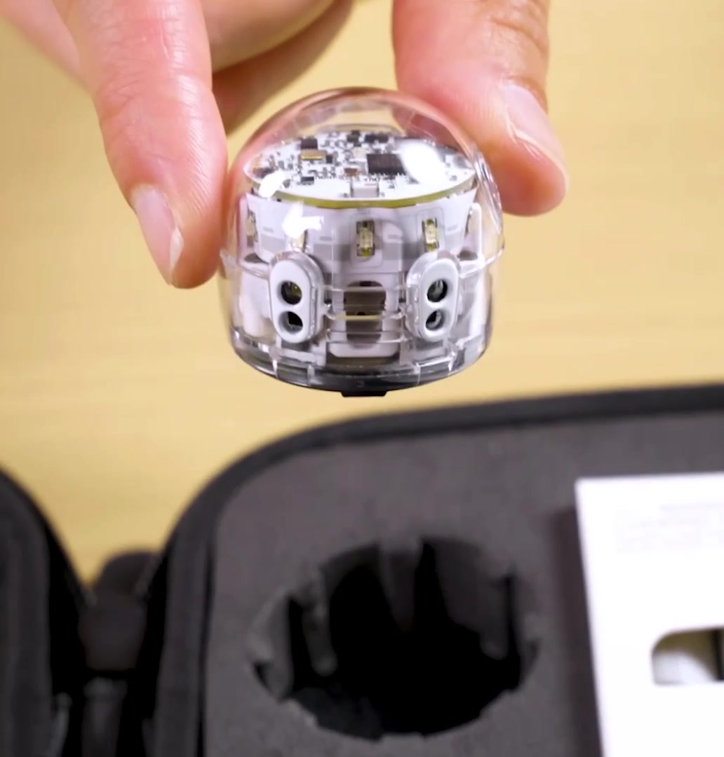 A hand lifting the Evo robot out of its zip case on a light wooden desk, close enough to see the circuit board through its clear shell.