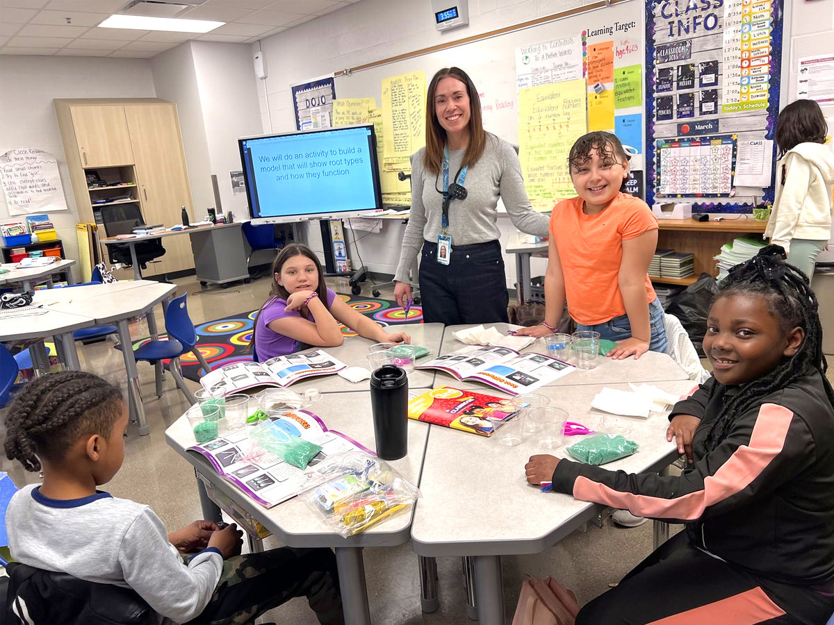 A Central Creativity instructor working with a small group at a table in a school classroom, the class teacher alongside them and the room plainly an ordinary school room.