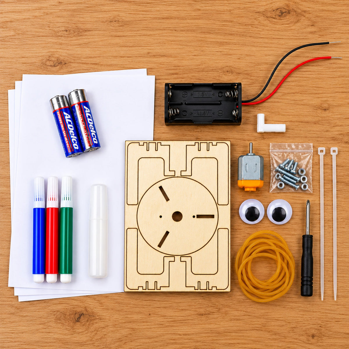 Everything in the Art Bot kit laid out on a desk: the laser-cut plywood board, the motor, a wired battery case, two AA batteries, three markers, screws and nuts, zip ties, rubber bands, two adhesive eyes, a Phillips screwdriver and sheets of drawing paper.