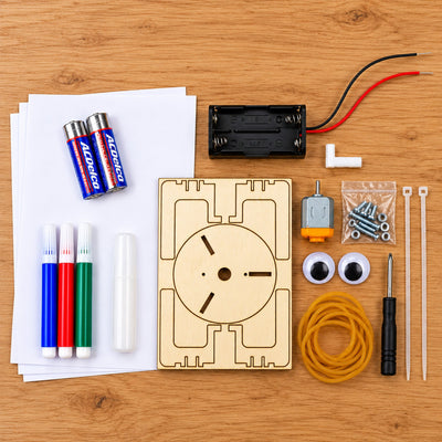 Everything in the Art Bot kit laid out on a desk: the laser-cut plywood board, the motor, a wired battery case, two AA batteries, three markers, screws and nuts, zip ties, rubber bands, two adhesive eyes, a Phillips screwdriver and sheets of drawing paper.