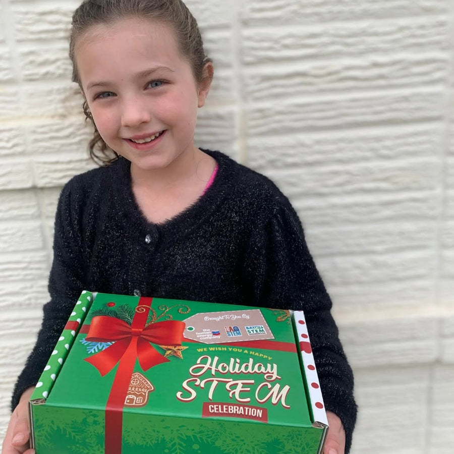 A girl holds the Holiday STEM Celebration box in front of a white brick wall.