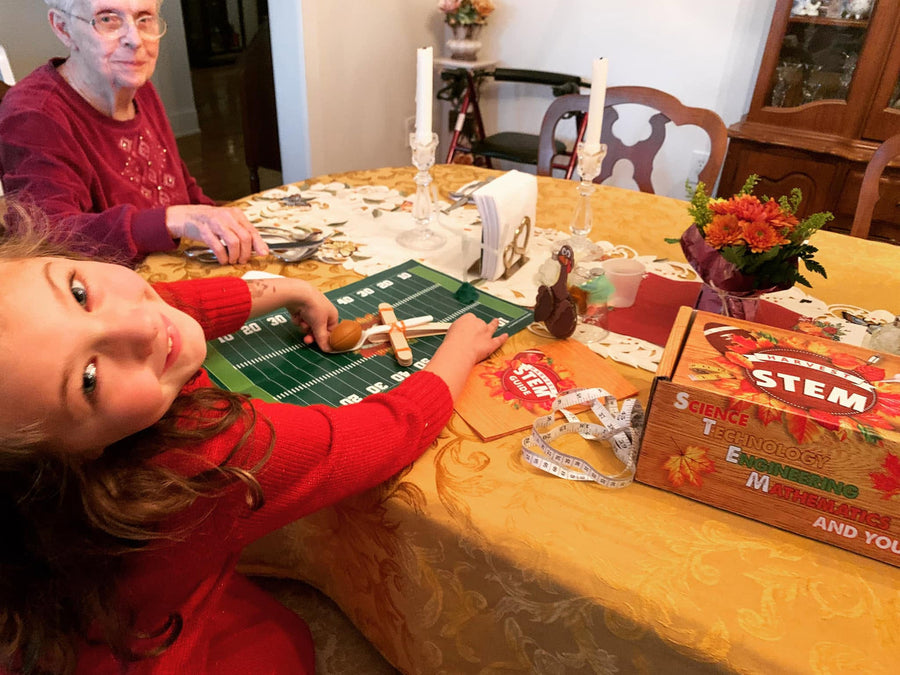 A girl in a red sweater adjusts a clothespin catapult on a paper football field at the dining table while her grandmother watches from across the table.