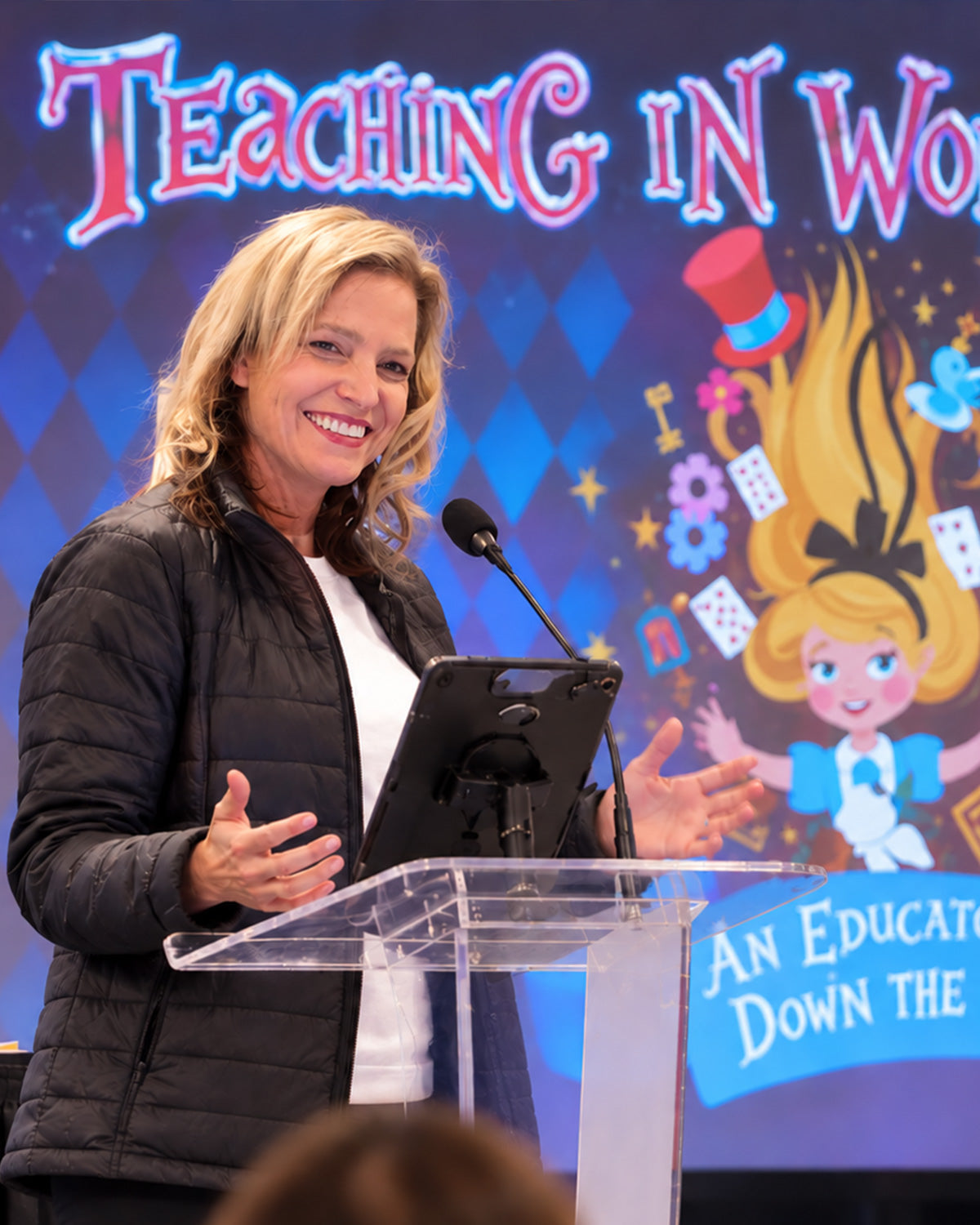 Jenny McLeod speaking at a clear lectern with a microphone, hands raised mid-sentence, in front of a large illustrated conference backdrop.