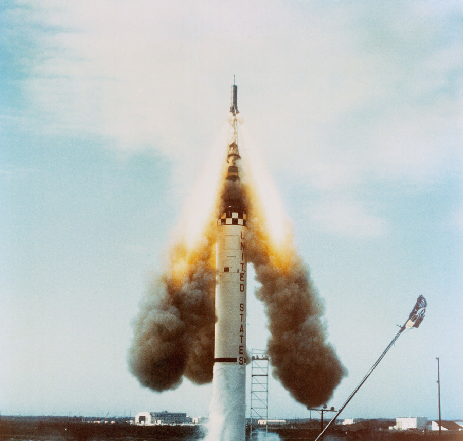 A Mercury-Redstone rocket lifting off, its service tower alongside and exhaust streaming across the pad