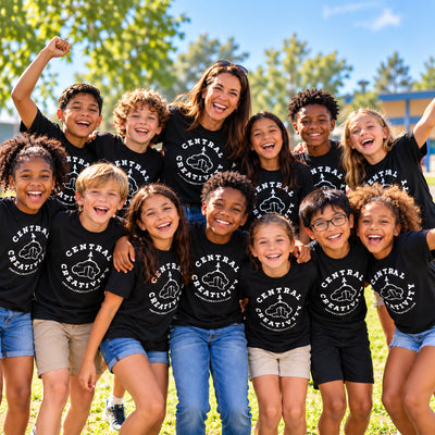 Thirteen children and their teacher outdoors in black Central Creativity t-shirts, crowded together laughing with their arms raised.