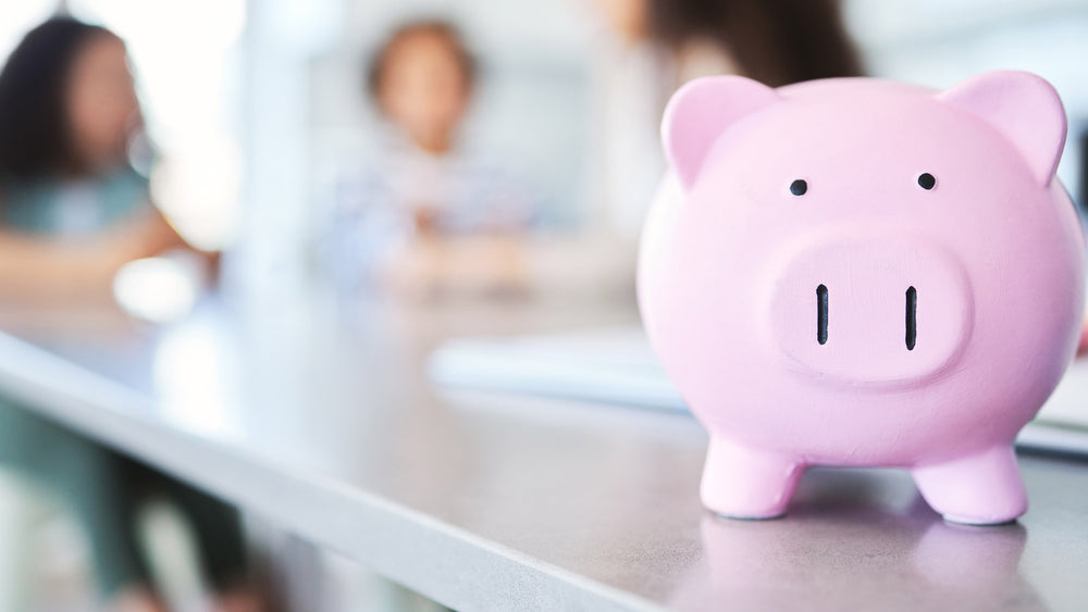 A pink piggy bank sits on a classroom counter with students working out of focus in the background.