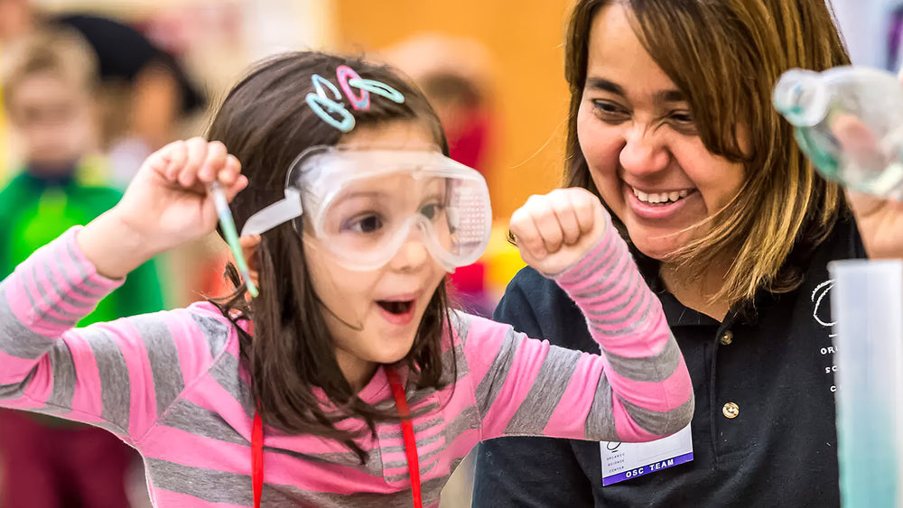 A young girl in safety goggles cheers with both fists raised while a smiling educator beside her holds up a bottle of bubbling liquid.