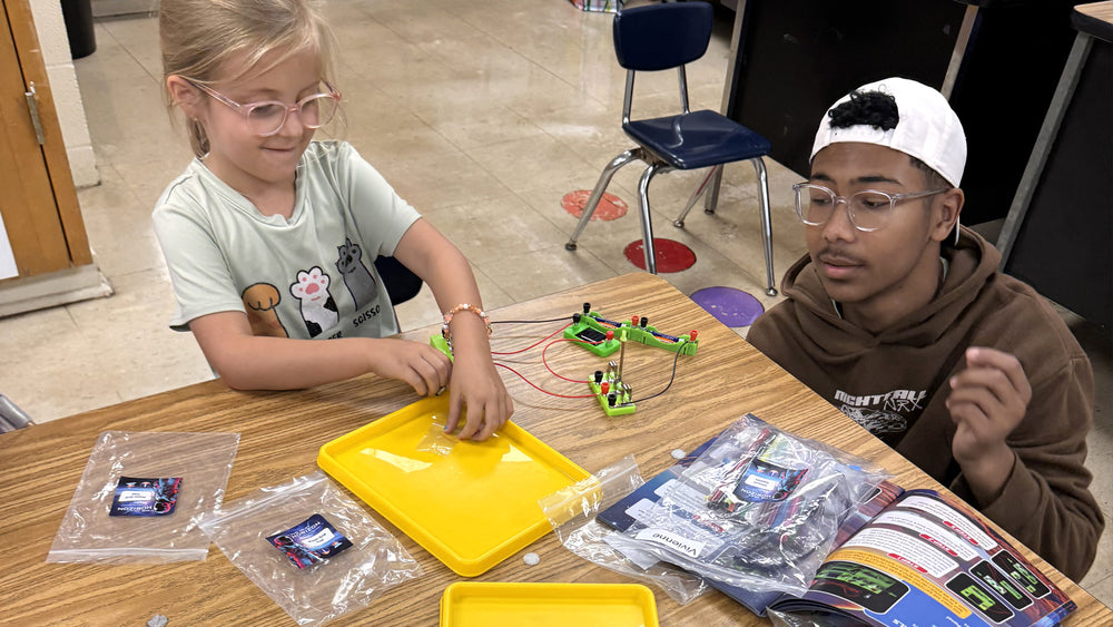 A younger student and an older student sit at the same classroom table building a snap-circuit project together, each working from the same kit.
