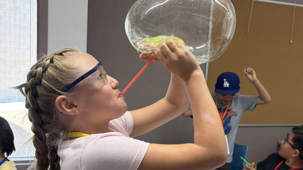 A girl in safety glasses blows a giant soap bubble through a straw while classmates work at tables behind her.