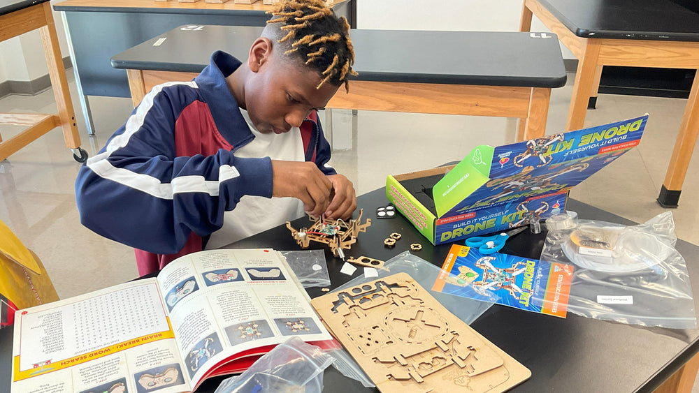 A student assembles a build-your-own drone at a classroom lab table, with the open kit box, illustrated instruction booklet, laser-cut parts and sealed component bags all laid out in front of him.